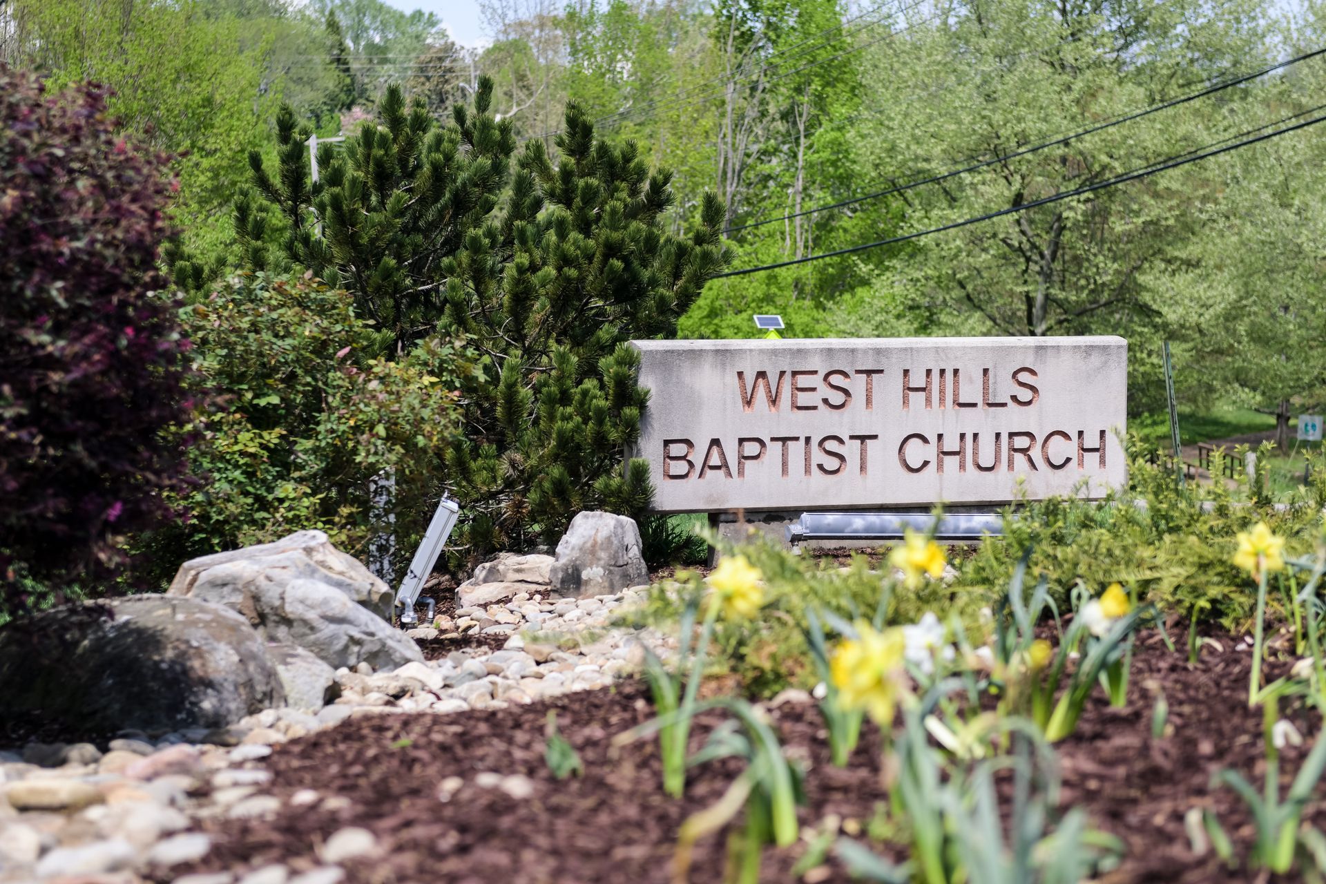 Sign for West Hills Baptist Church in a garden setting, surrounded by plants and flowers.