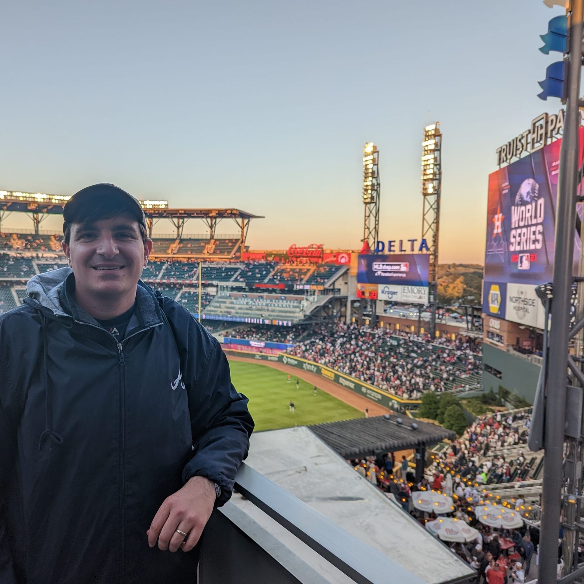 Man at a baseball stadium at sunset, looking at the field and the crowd. The scoreboard displays 