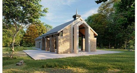 A modern stone building with a pointed roof sits in a grassy clearing surrounded by trees under a blue sky.