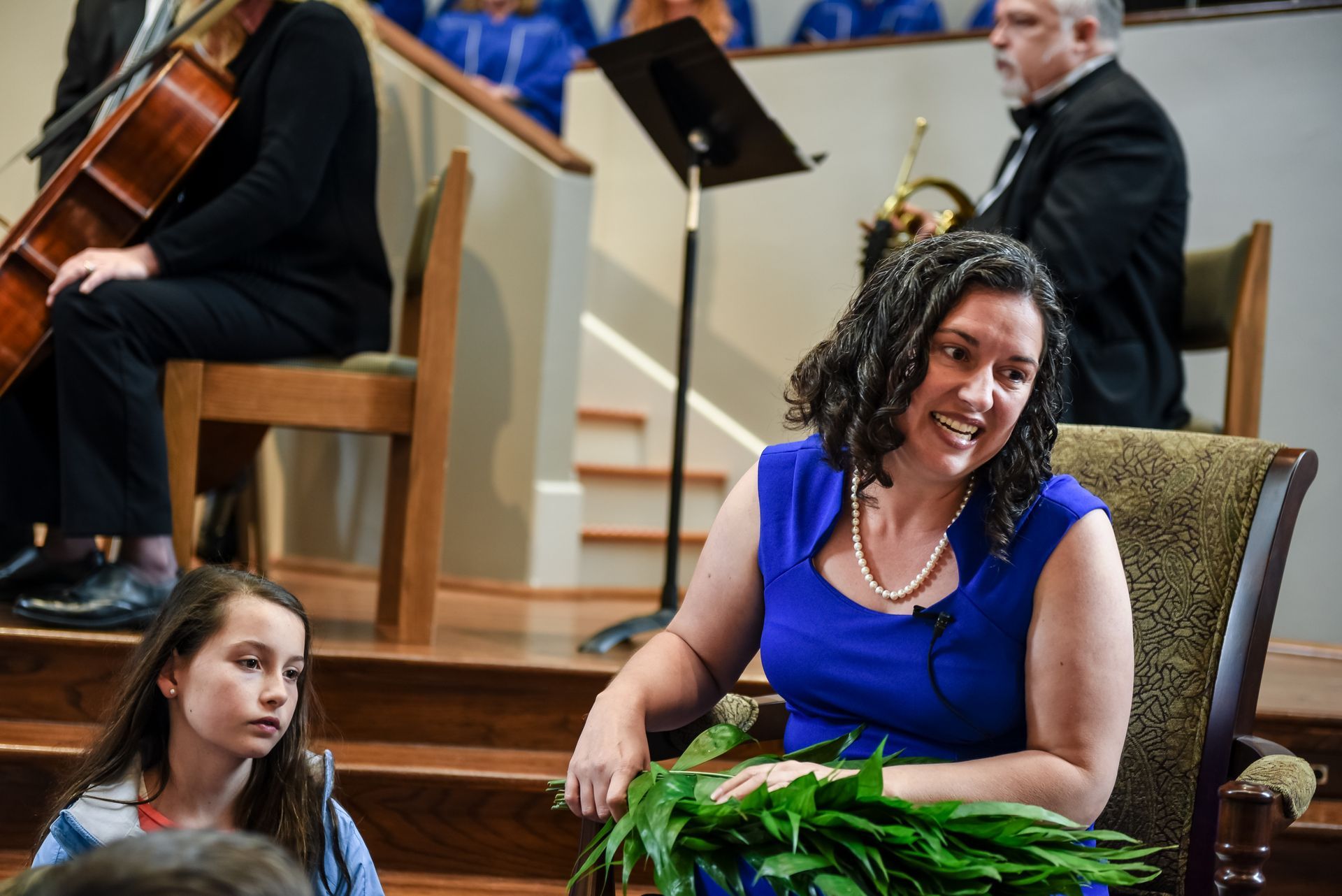 A woman in a blue dress holds greenery, smiling, talking to a young girl. Musicians and a choir are in a church setting.
