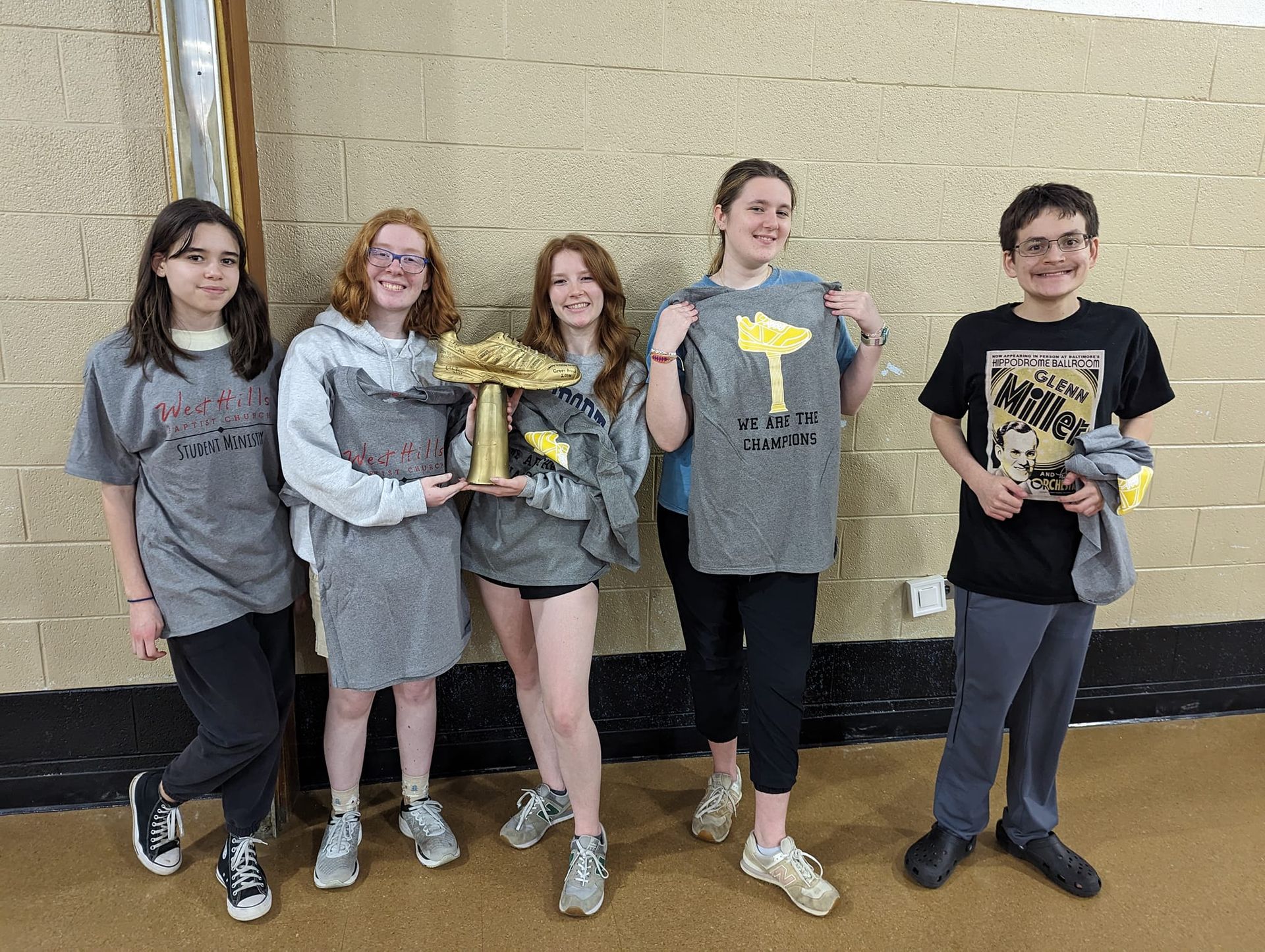 Five teens, two holding a trophy and another holding up a shirt. They stand against a beige wall, smiling, with casual clothes.