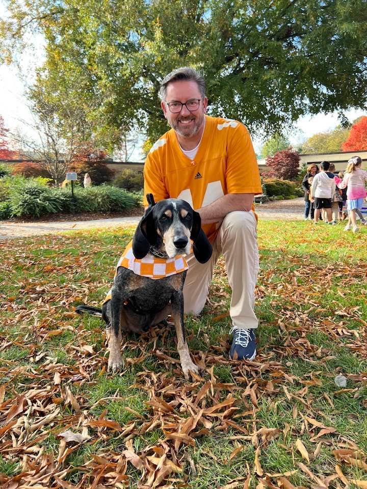 A man in an orange shirt kneels beside a dog wearing a matching orange collar. They are outside in a grassy area.