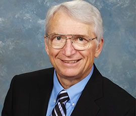 Smiling older man with glasses, wearing a suit and tie, against a blurred blue background.