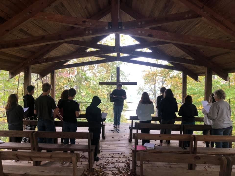 Group of people singing in front of a wooden cross in an outdoor gazebo. Surrounded by trees, it's likely autumn.