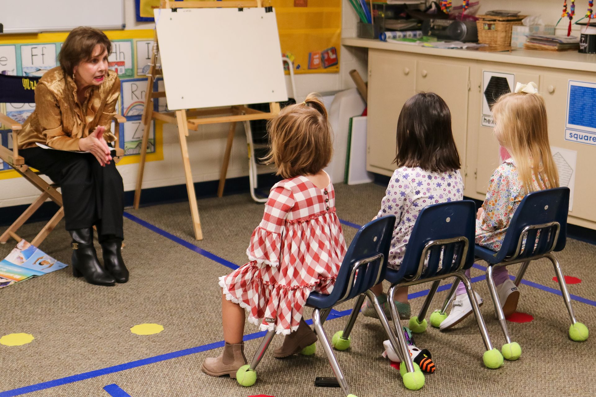 A teacher sits facing three young children in a classroom, gesturing towards a blank whiteboard.