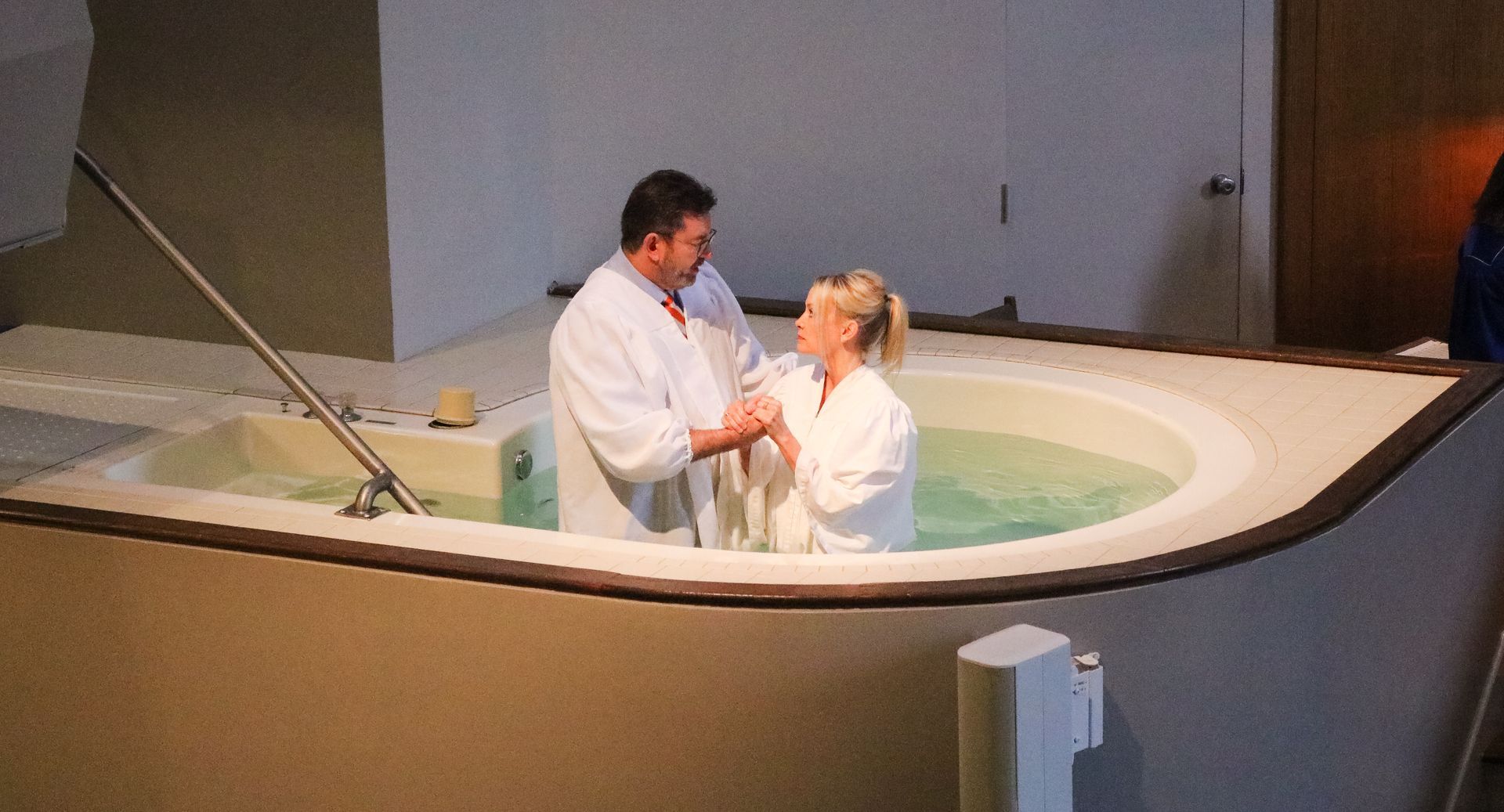 A man in a white robe baptizes a woman in a pool inside a church. Both are smiling, holding hands, in chest-high water.