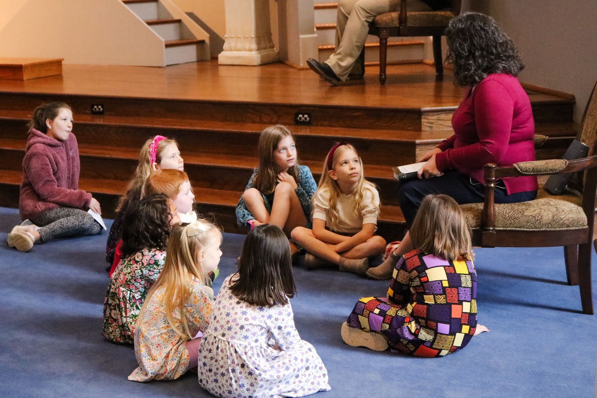A woman reads to a group of young girls seated on a blue carpet. They are in a room with a stage and wooden stairs.