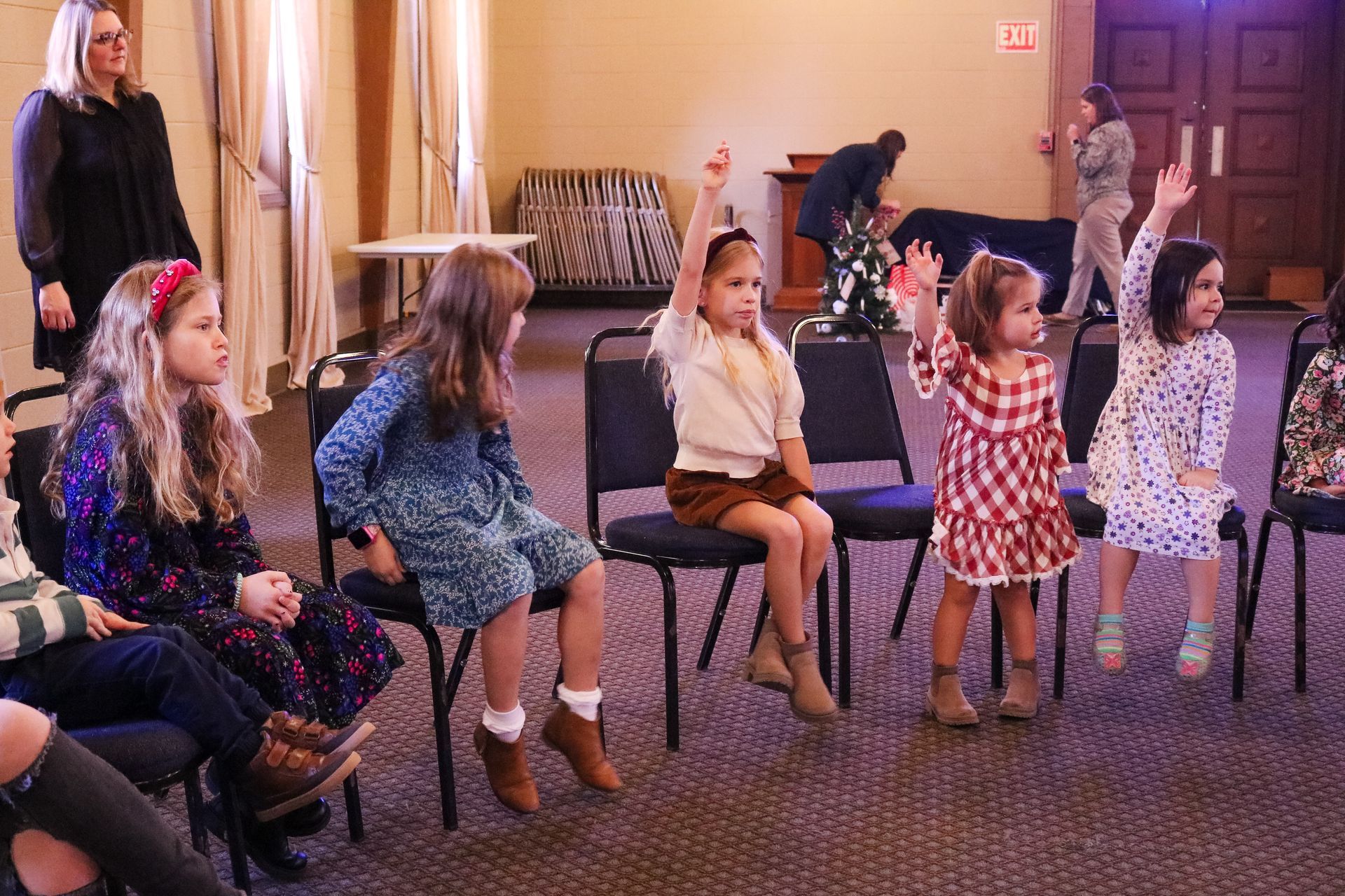Children seated in chairs raise their hands. A woman watches them, likely a teacher, inside a room.