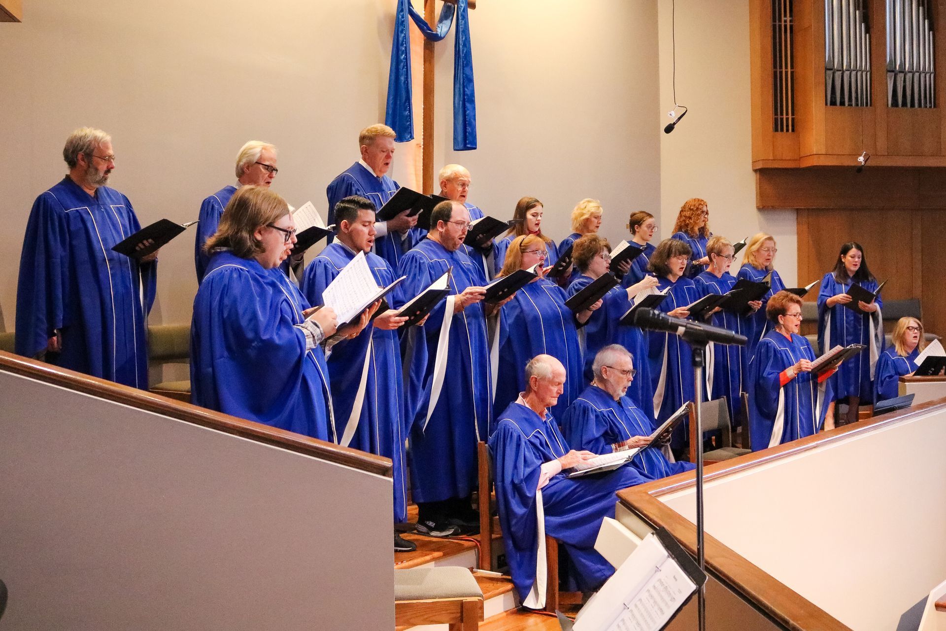 A choir in blue robes sings from a church balcony, holding music sheets. A blue cross hangs on the wall behind them.