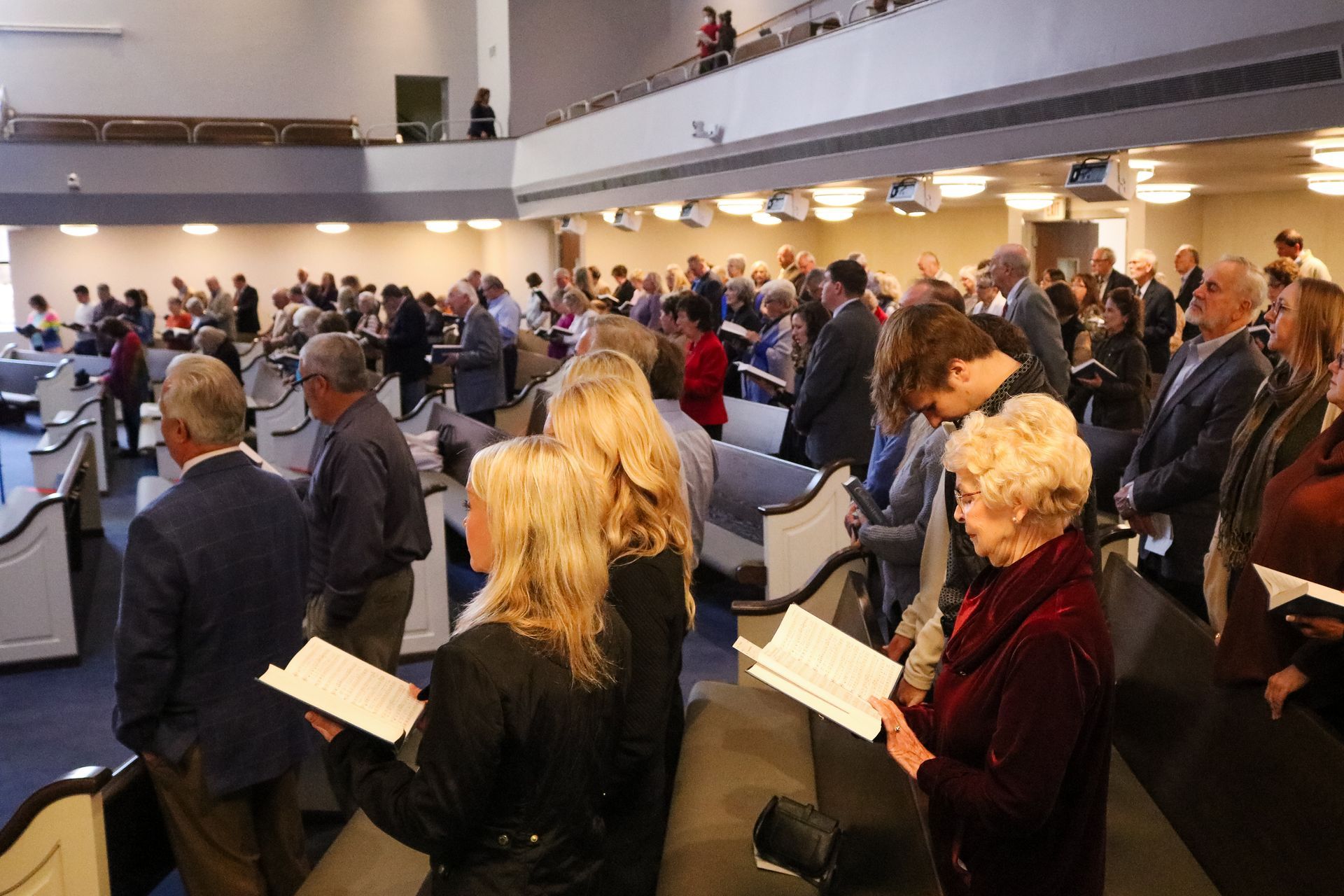 People stand and sing in a light-filled church sanctuary, holding songbooks, with a balcony overhead.