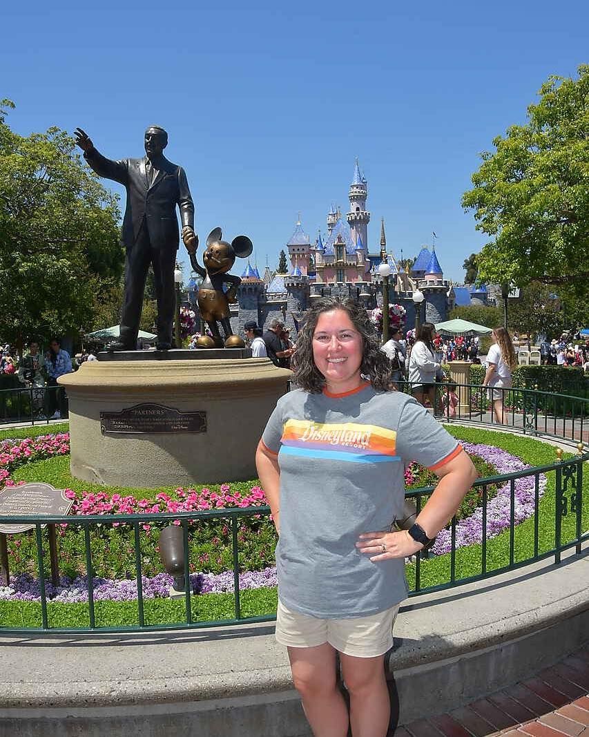 Woman in front of the Partners statue and Sleeping Beauty Castle at Disneyland. She wears a gray t-shirt and khaki shorts, with a smile and hands on hips.