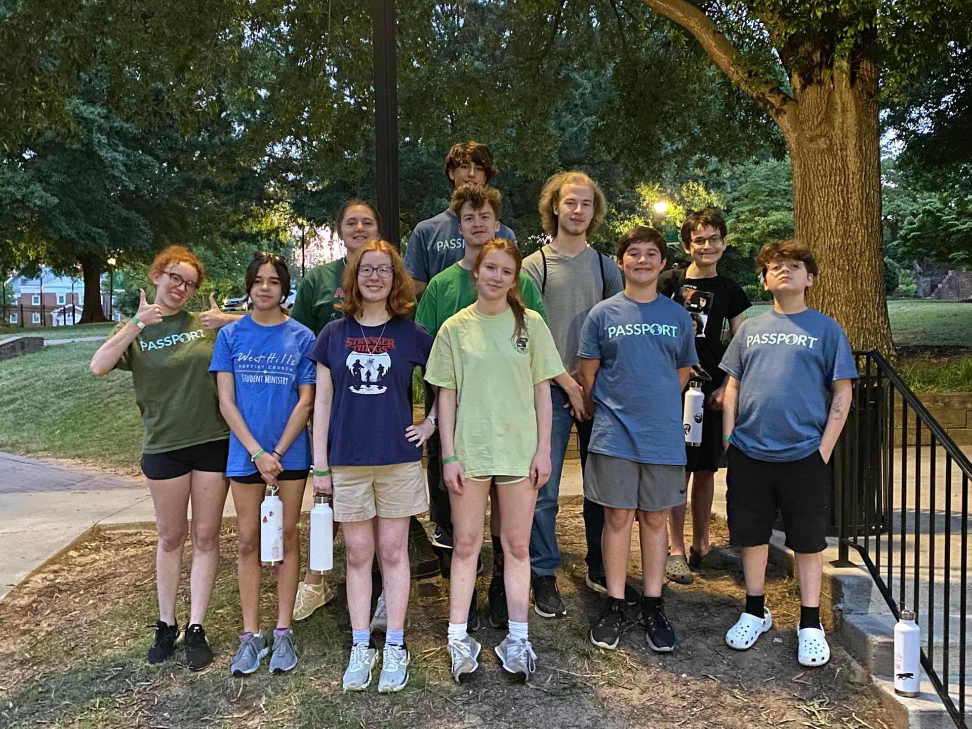 A group of teens stands together outside, some holding water bottles. They are of varying heights and wear casual clothes.