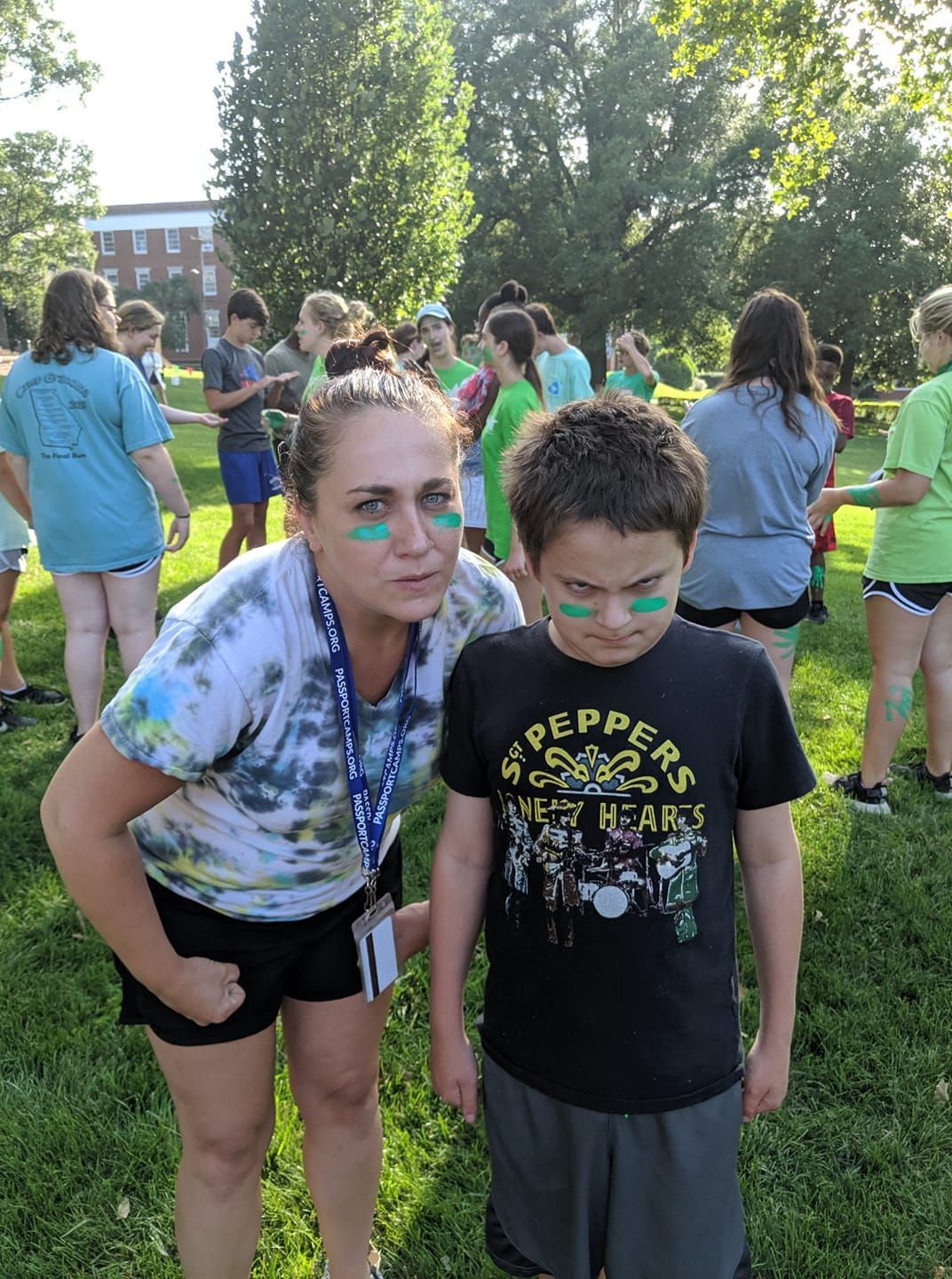 Woman and boy with green face paint stand in a park with a group of people. They are wearing t-shirts and shorts.