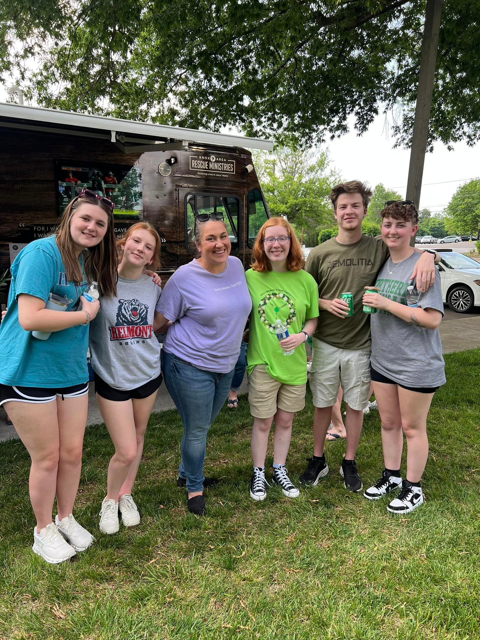 Six young people smiling and posing together in front of a food truck, outdoors on a grassy area.