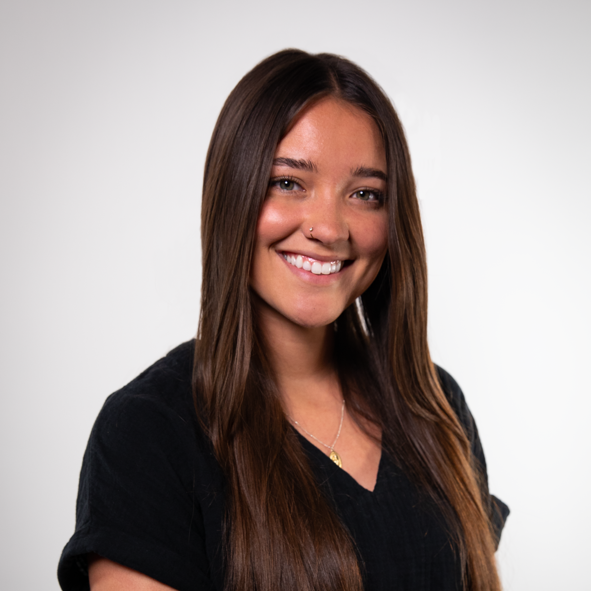 Woman with long brown hair smiling at the camera, wearing a black v-neck shirt, and a nose ring. White background.