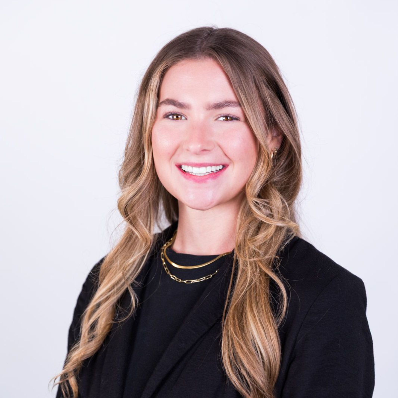 Woman with long wavy hair smiles, wearing a black blazer and gold necklace.