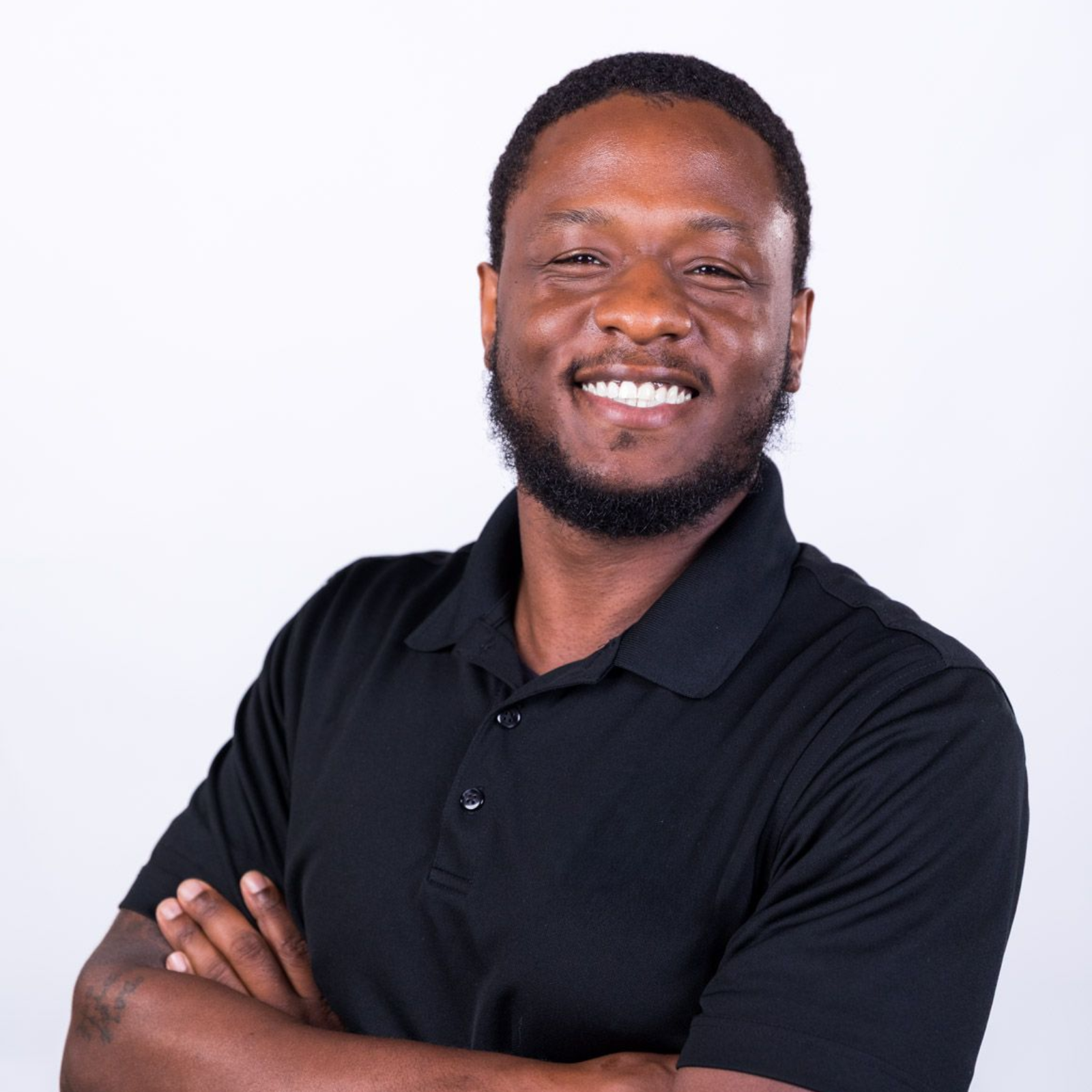 Man with crossed arms smiling; wearing a black polo shirt, against a white backdrop.