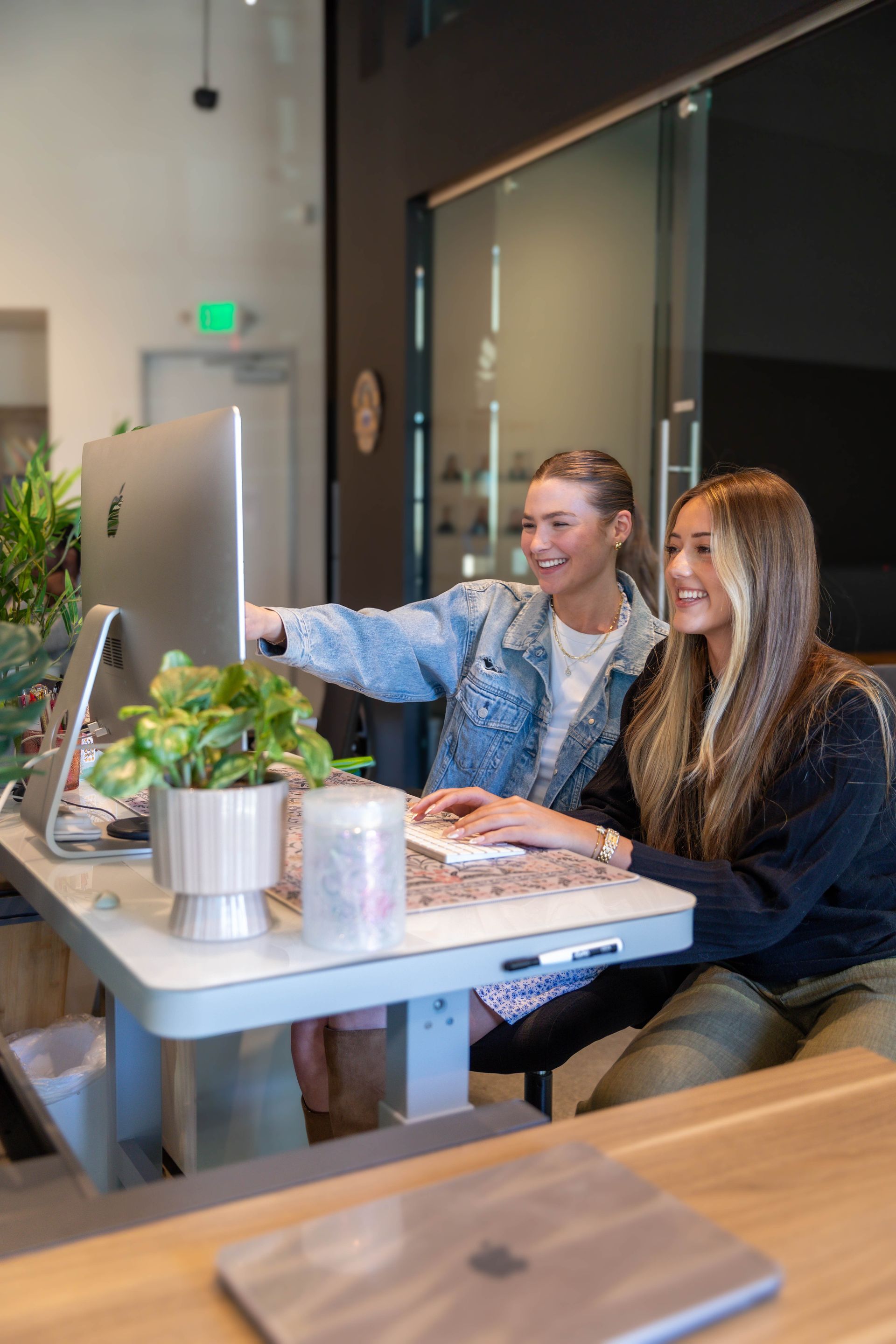 Two people smiling at a laptop in a modern café, seated at a table with a plant and drinks.