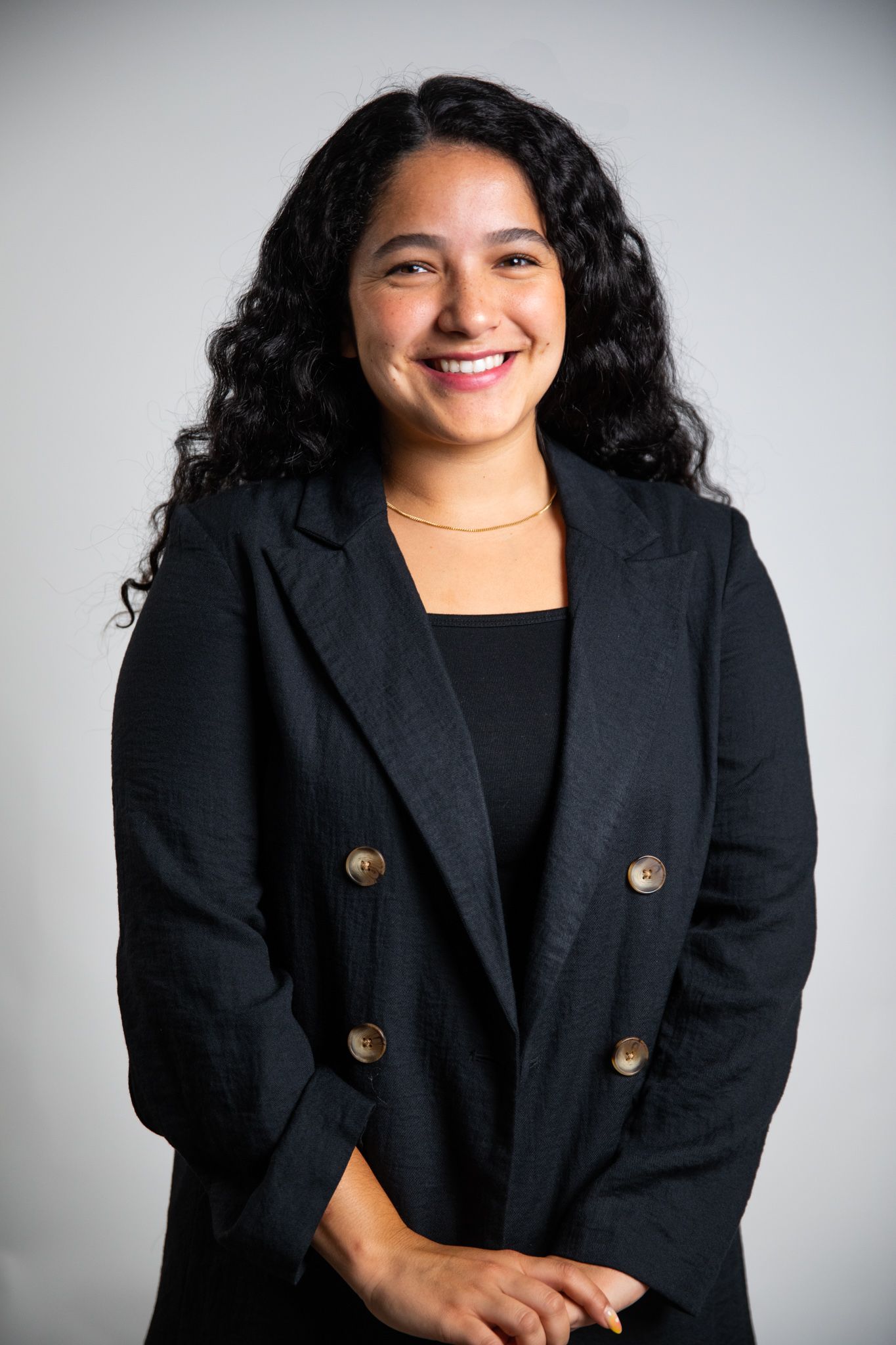 A woman with curly hair is wearing a black jacket and smiling.