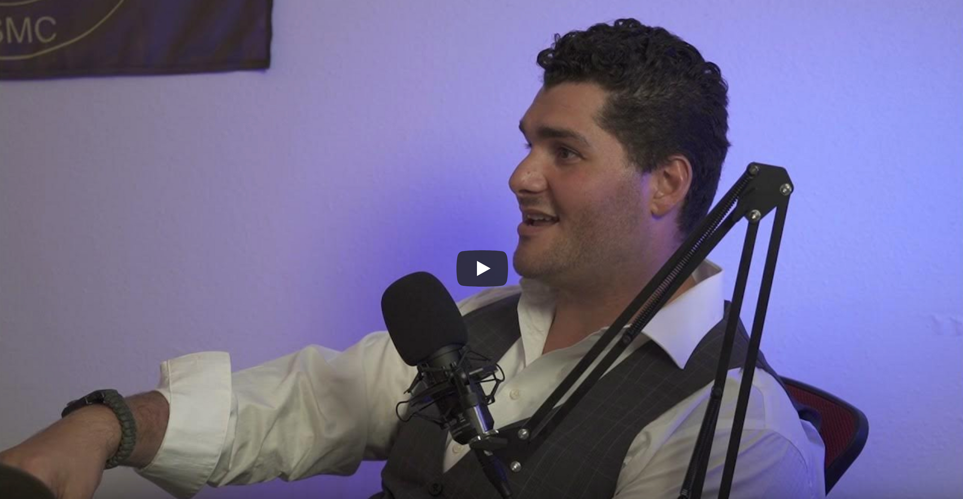 A man is sitting in front of a microphone with a play button on the screen.