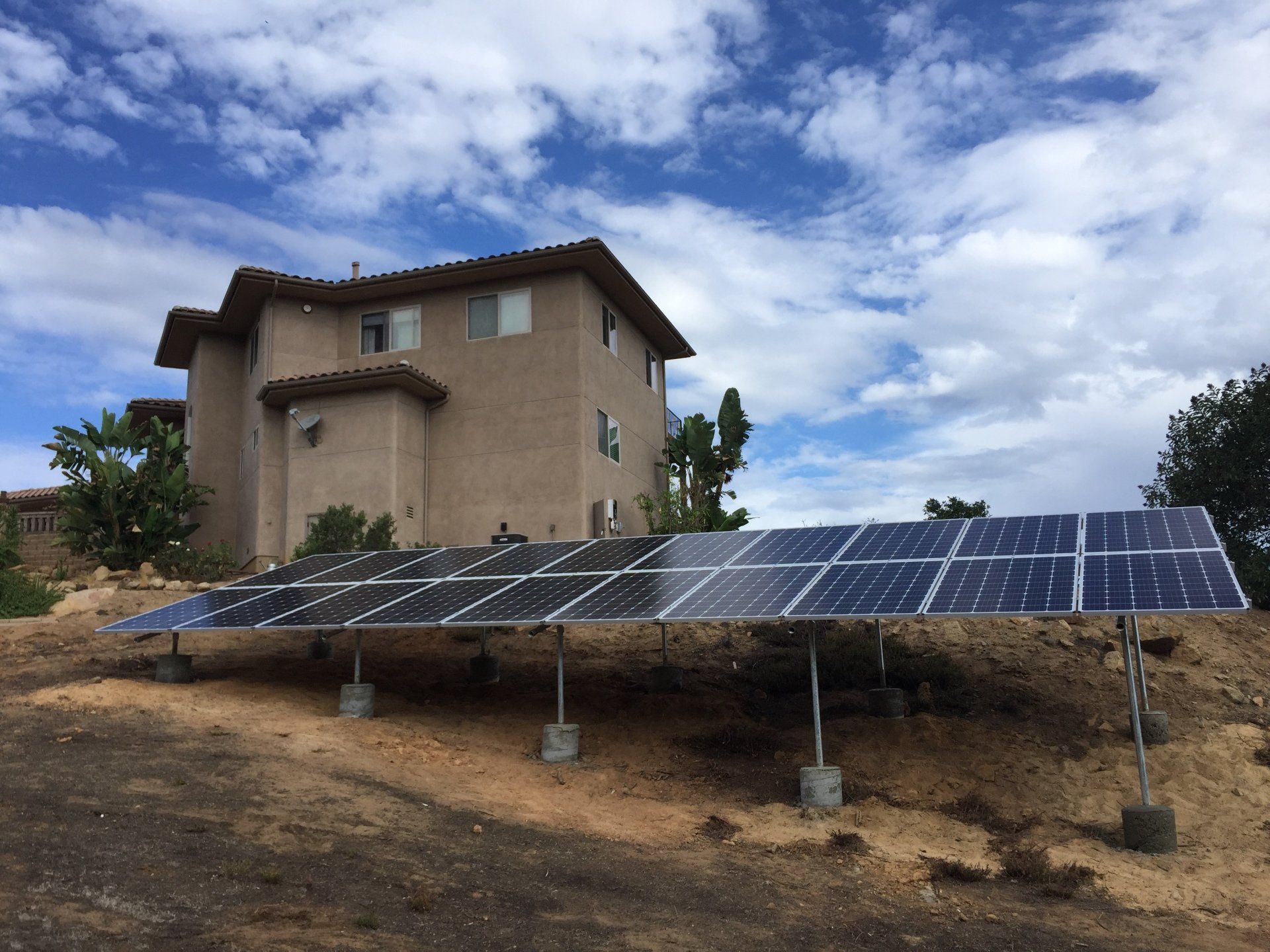 Solar panels on a dirt lot beside a beige house under a partly cloudy sky