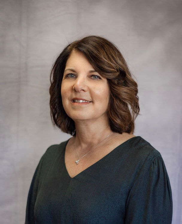 Woman with short, dark wavy hair in a dark green v-neck top smiles at the camera against a gray backdrop.