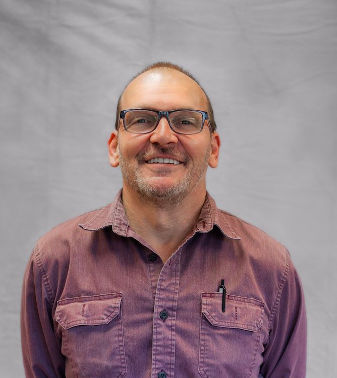 Man wearing glasses and a maroon button-up shirt smiles in front of a gray backdrop.
