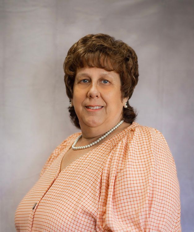 Woman with curly brown hair, wearing a patterned orange shirt and pearl necklace, smiling.
