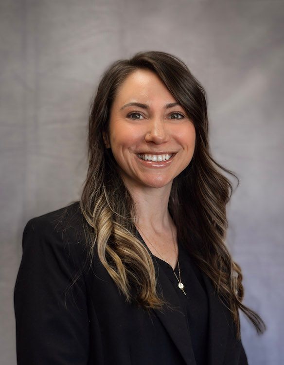 Woman with brown hair smiles, wearing a black blazer against a gray background.