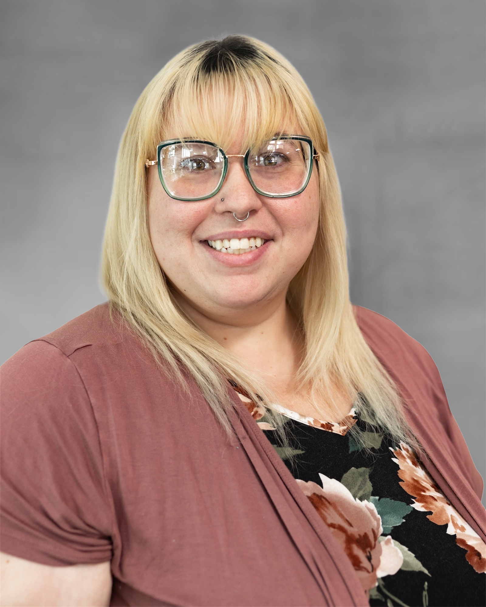 Woman with blonde hair, glasses, and a nose ring smiles, wearing a pink cardigan over a floral top.