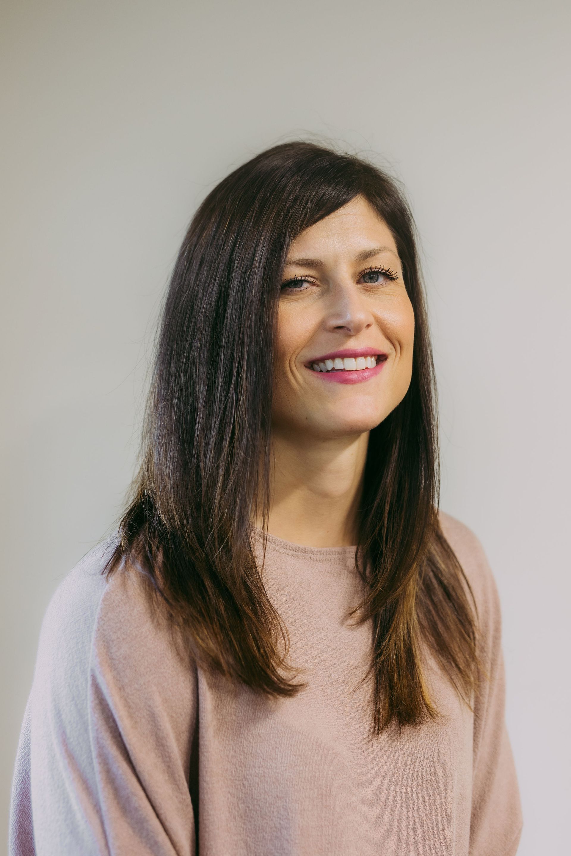Woman with dark hair smiles, wearing a pink sweater, standing in front of a neutral wall.