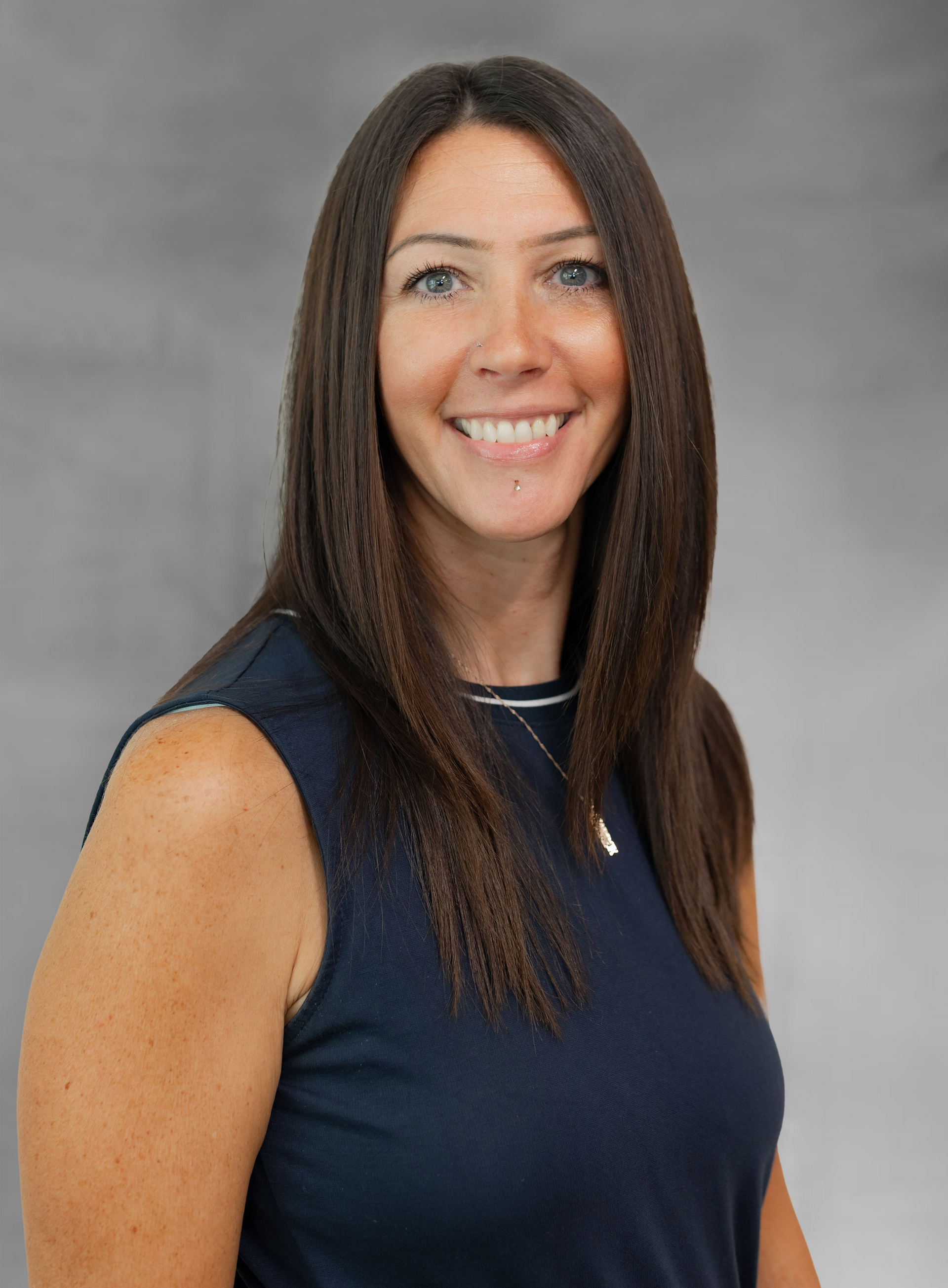 Woman with dark hair smiles, wearing a navy sleeveless top, standing in front of a gray backdrop.