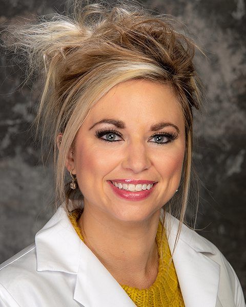 Woman with blonde updo smiles, wearing a yellow sweater and white lab coat.
