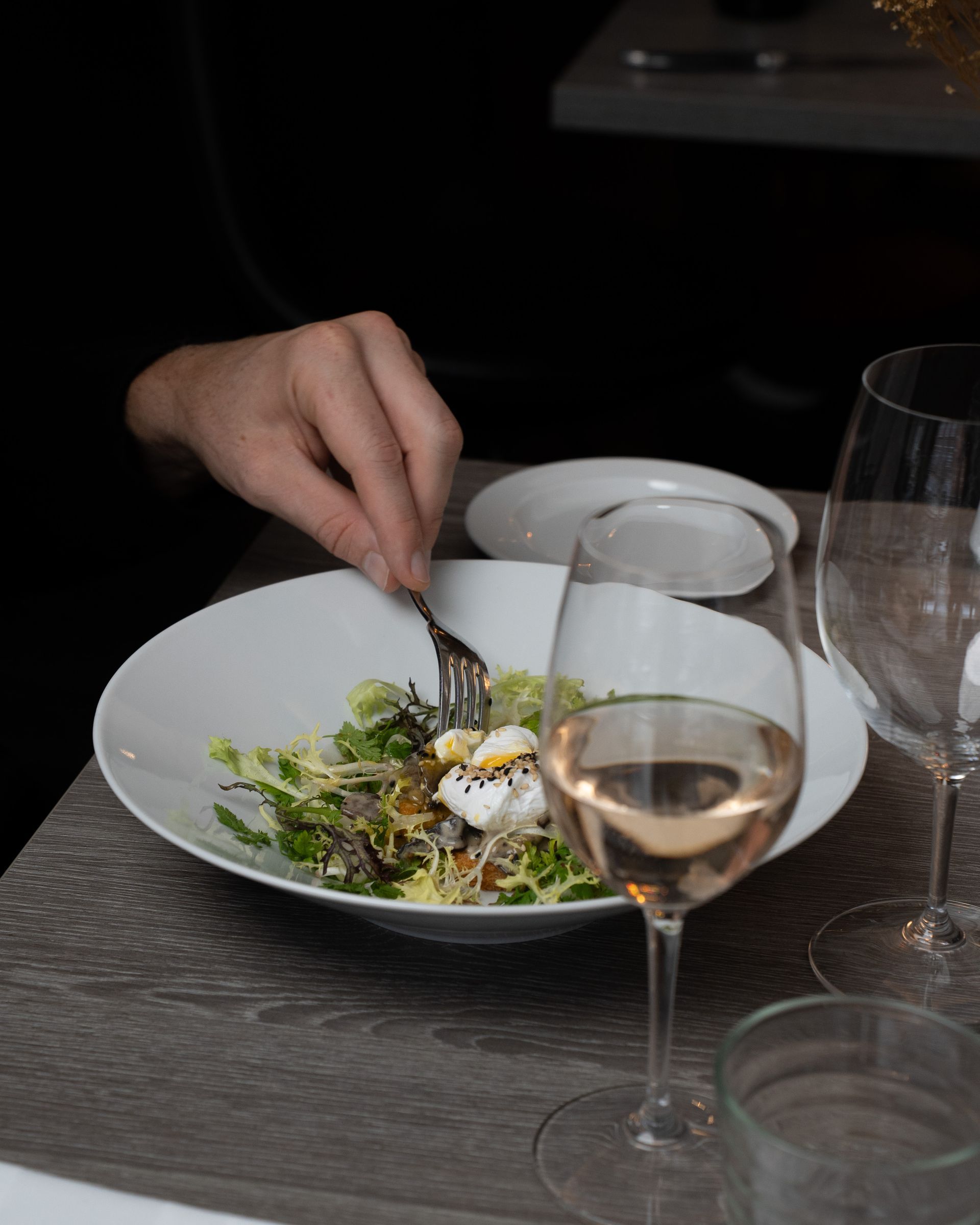 Person spiser salat med pocheret æg og rosévin ved et bord.