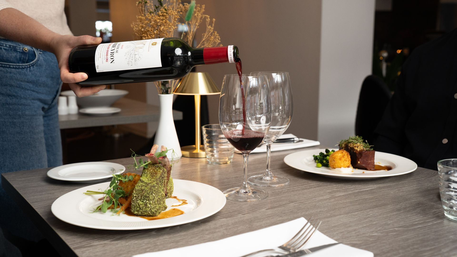 Nogen hælder rødvin i et glas ved et restaurantbord med tallerkener fyldt med mad.