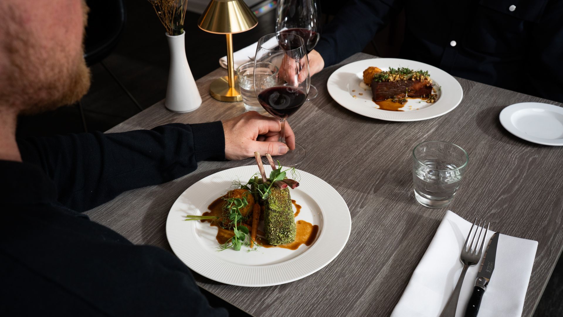 To personer ved et restaurantbord spiser på tallerkener med mad og drikker rødvin.