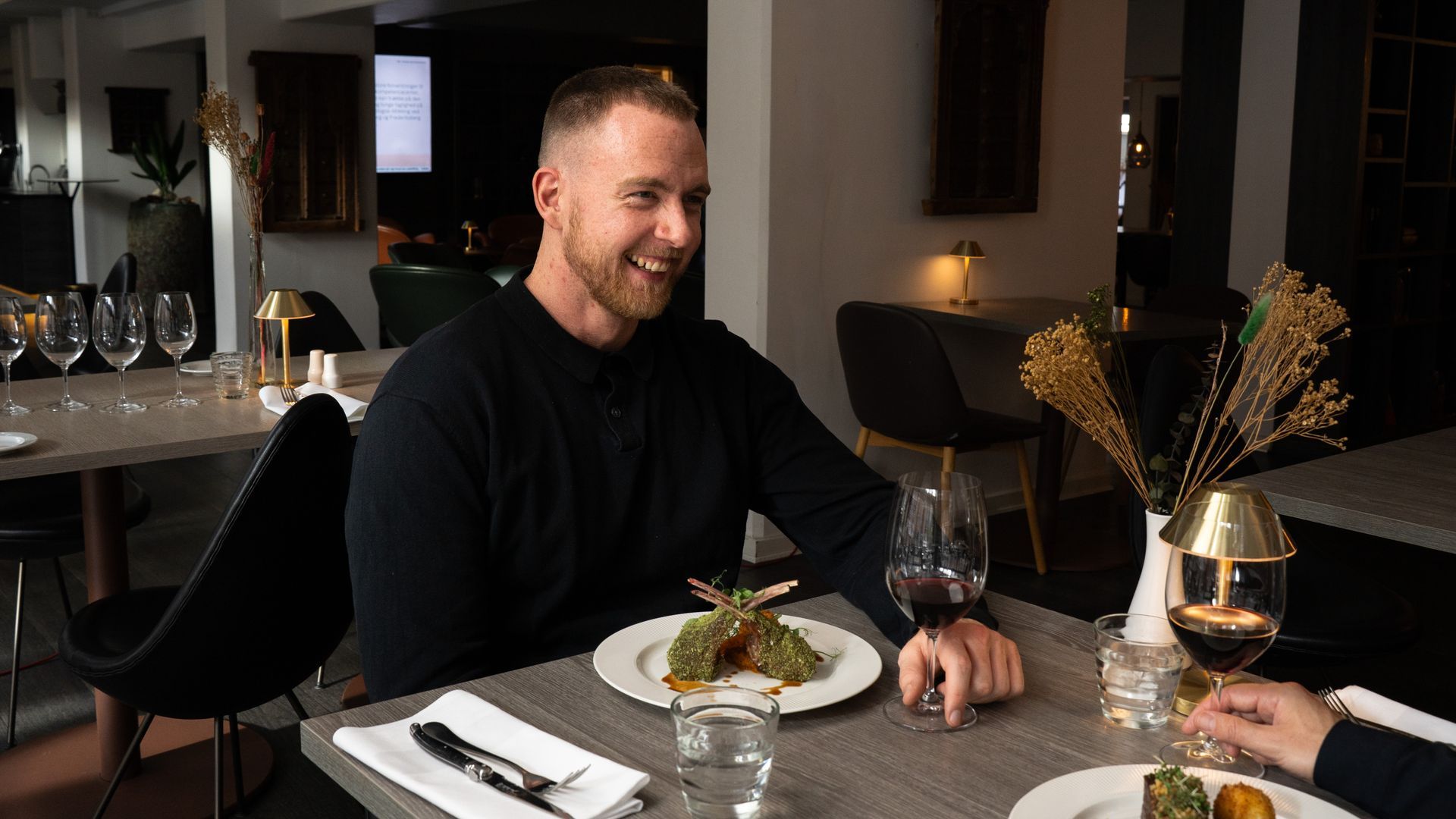 Mand griner ved et bord på en restaurant med mad og vin.