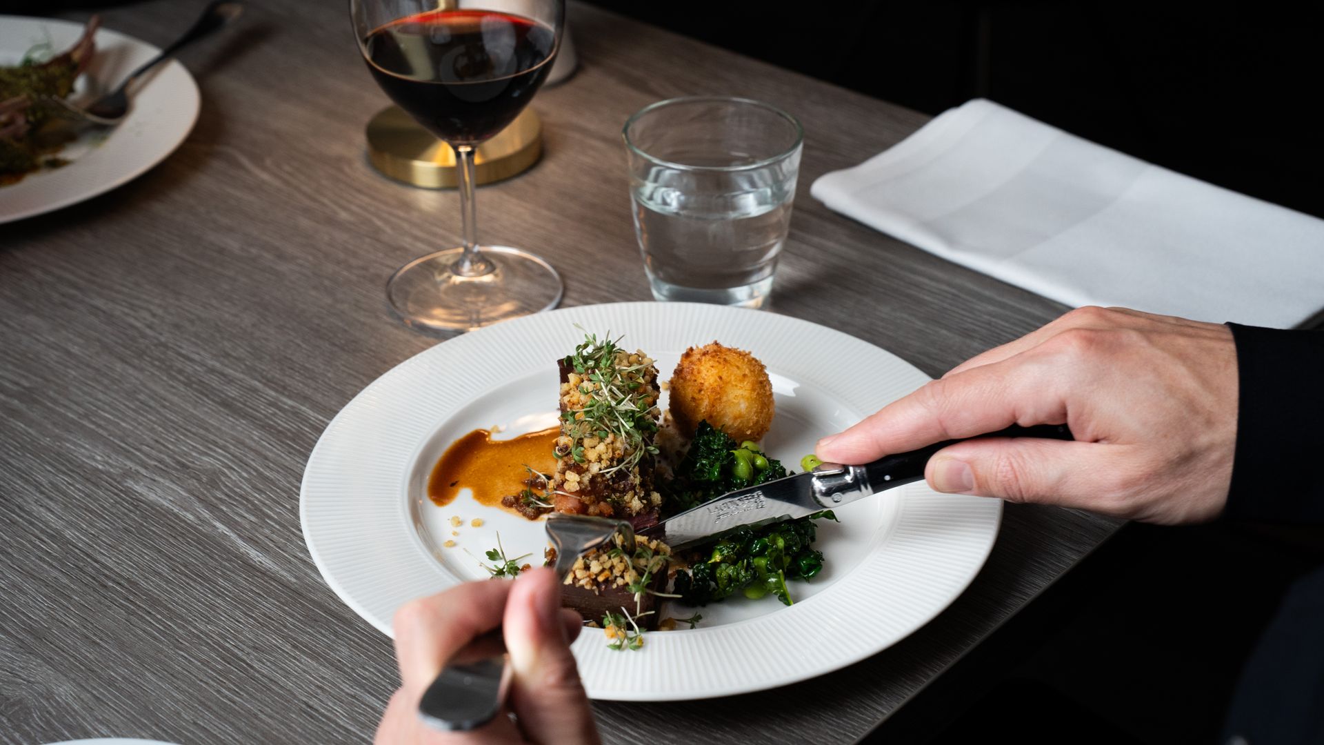 Person skærer kød på en hvid tallerken ved et restaurantbord med vin og vand.