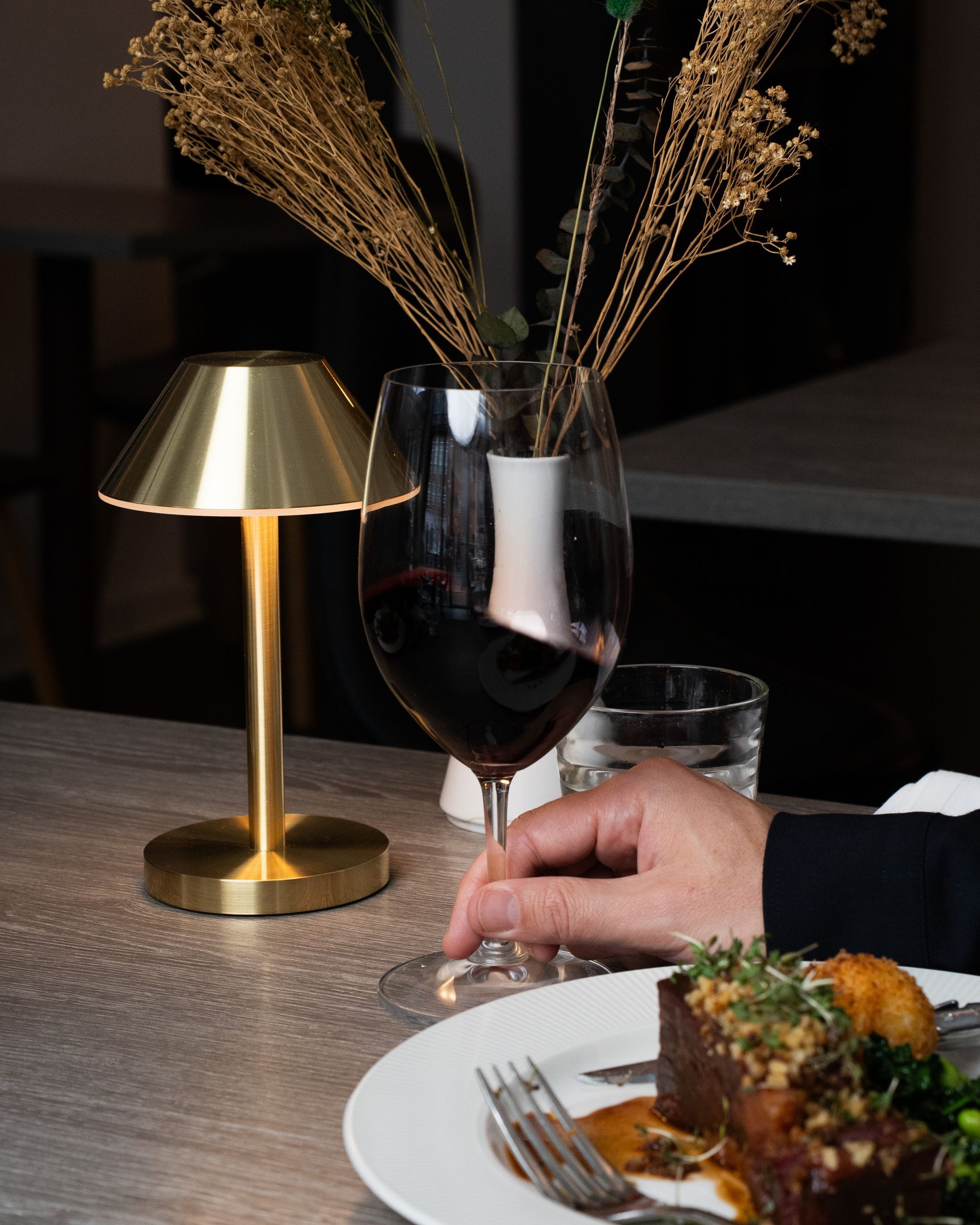 En gylden lampe oplyser et dækket bord til middag. En person holder et vinglas, og en tallerken med mad står i nærheden.