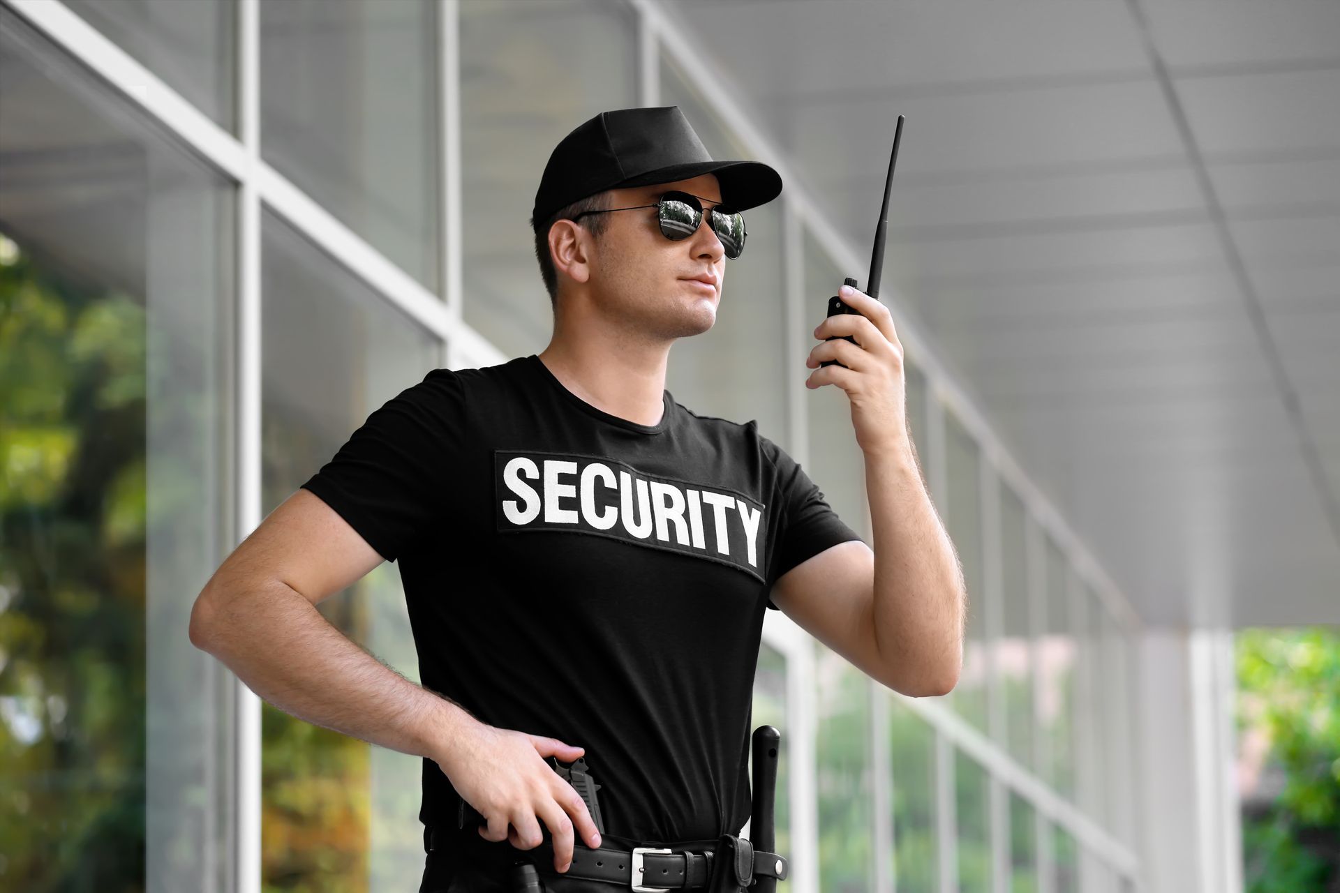 A security guard in a black uniform and sunglasses holds a walkie-talkie outside a building with glass walls. A security guard in a black uniform and sunglasses holds a walkie-talkie outside a building with glass walls.