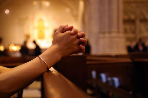 Hands clasped in prayer resting on a wooden church pew, with a blurred altar and interior in the background.