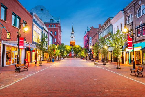 A brick-paved pedestrian street lined with historic red brick buildings, trees, and street lamps, leading to a church spire.