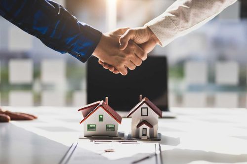 Two people shaking hands over two miniature model houses on a desk, representing a real estate deal.