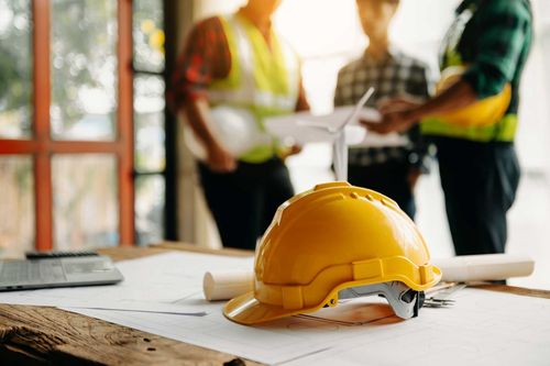 A yellow hard hat with a miniature wind turbine model sits on blueprints, with blurred construction workers behind.