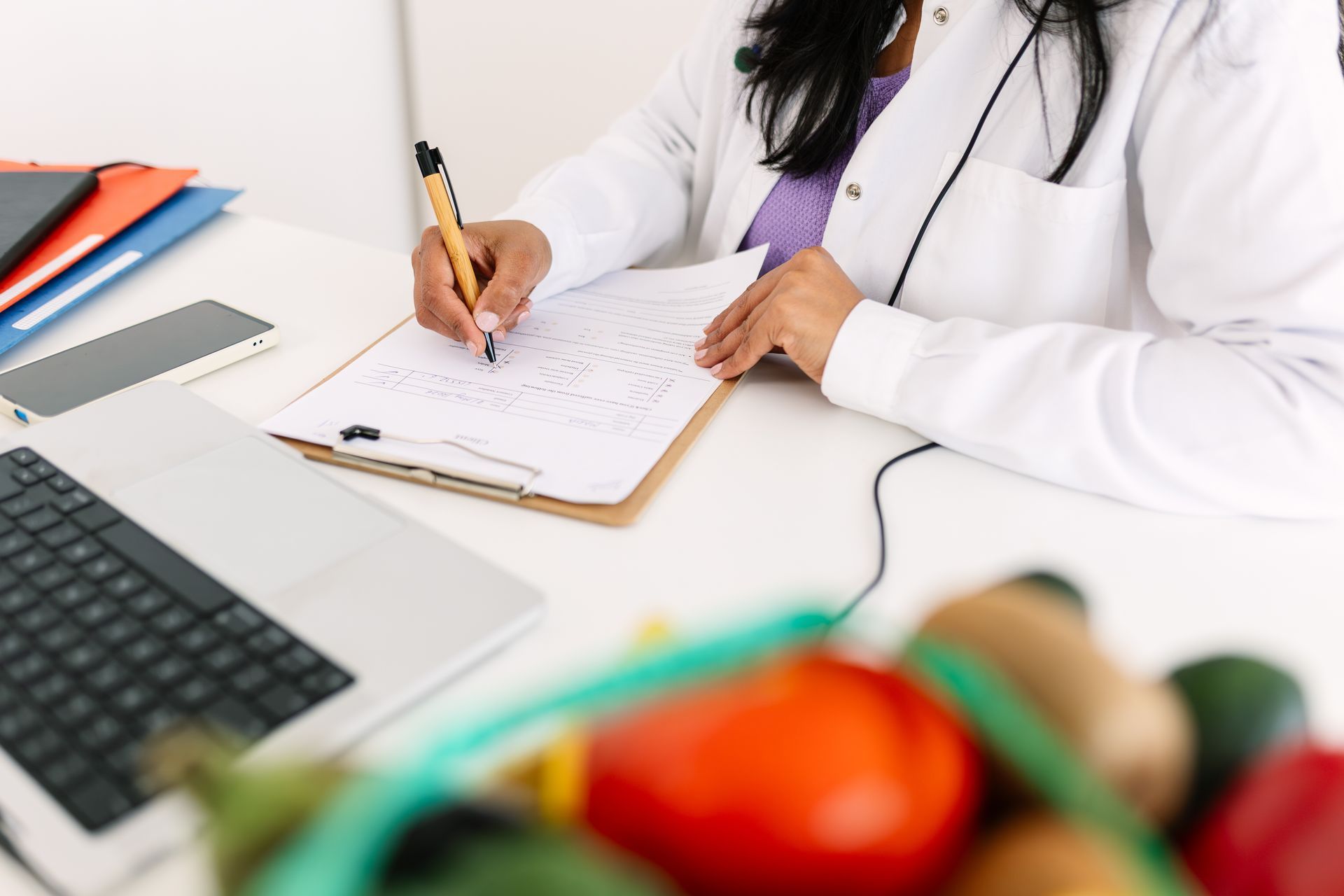 Doctor writing on clipboard beside a laptop, with medical tools in a clinic office.