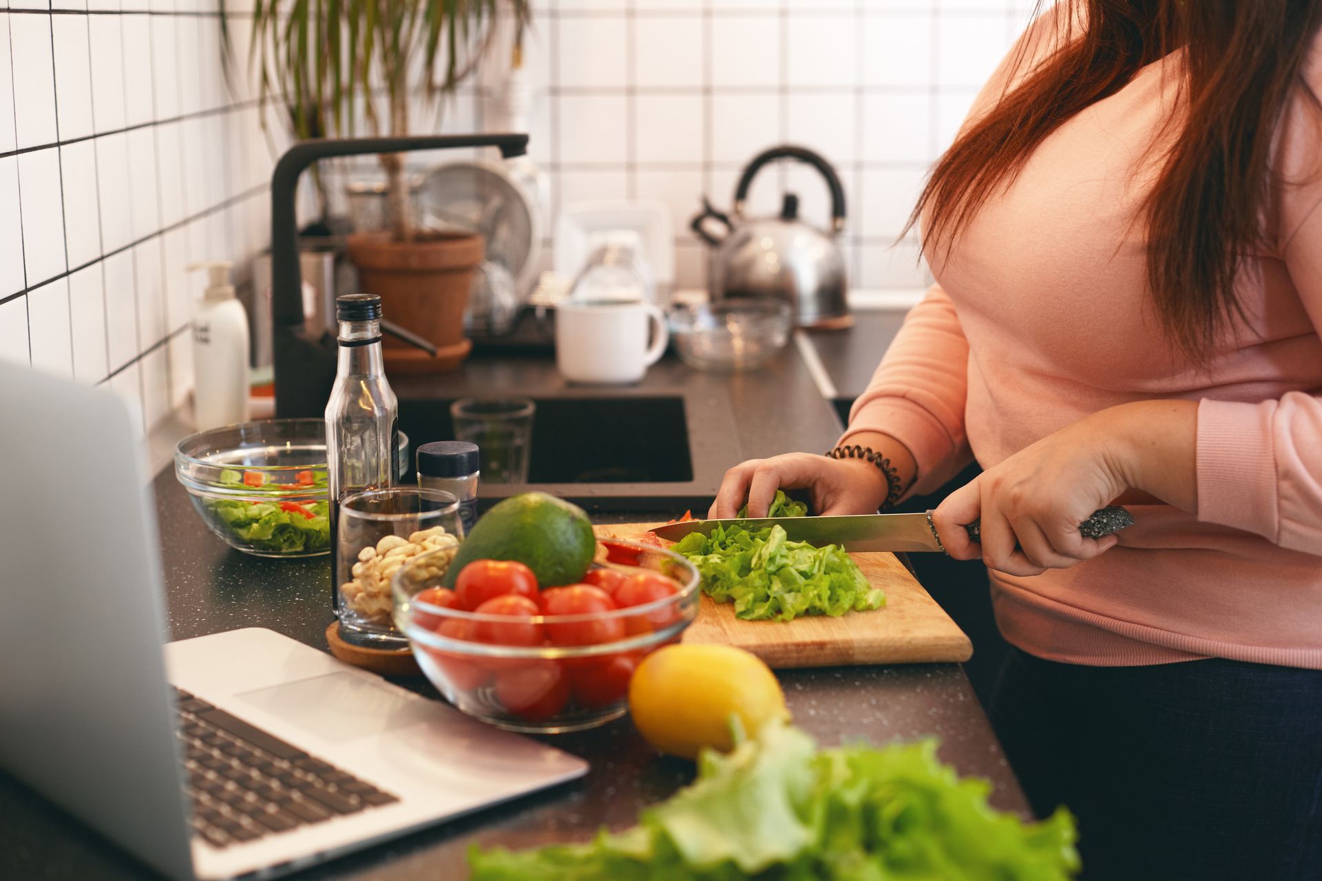 Person chopping vegetables in a kitchen beside a laptop, with salad ingredients on the counter.
