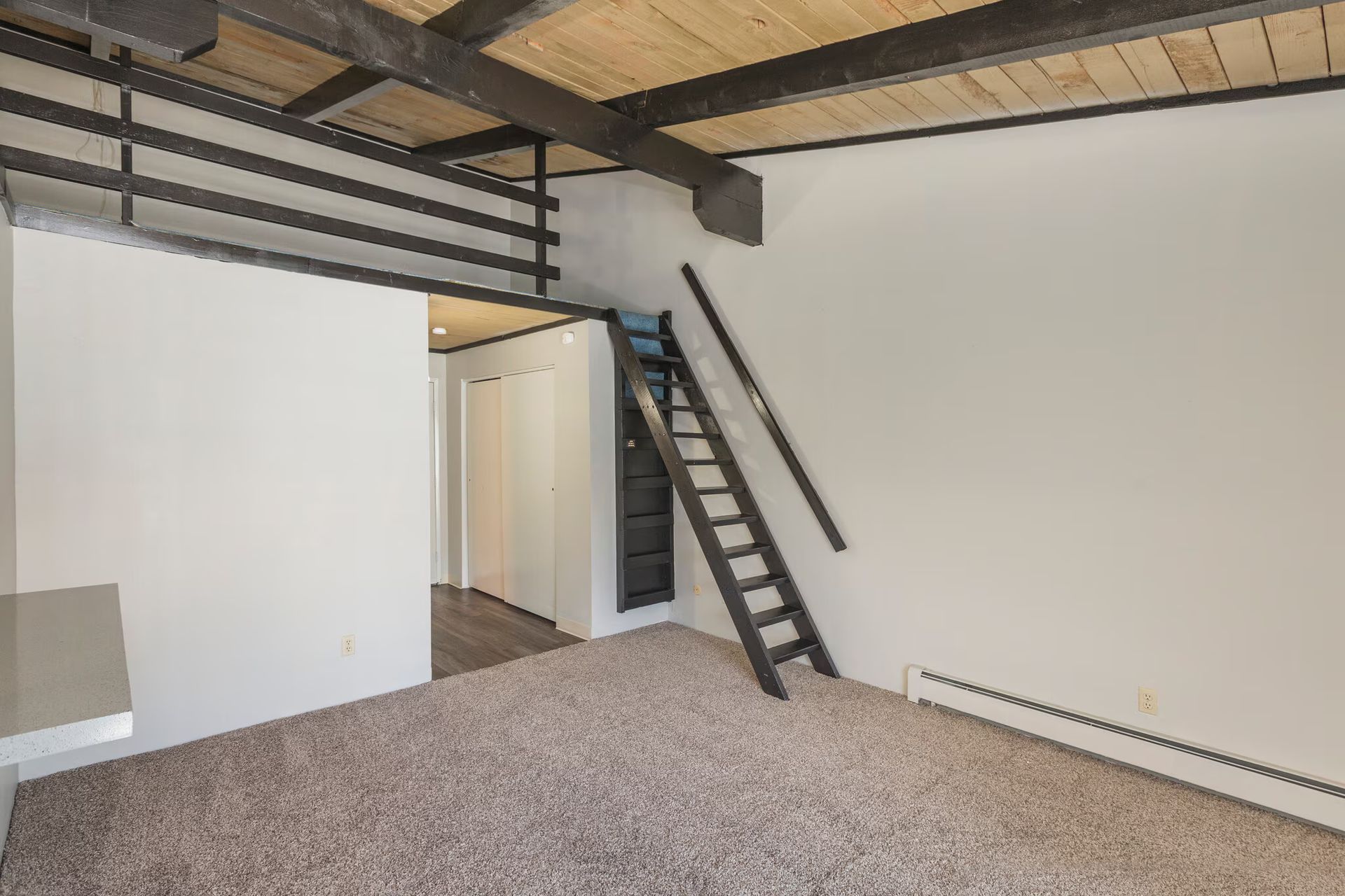 Interior of a room with a loft and dark wooden ladder. Beige carpet and white walls.