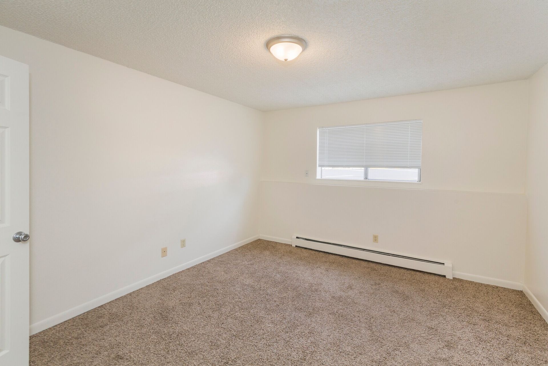 Empty bedroom with beige carpet, white walls, small window, and heater.