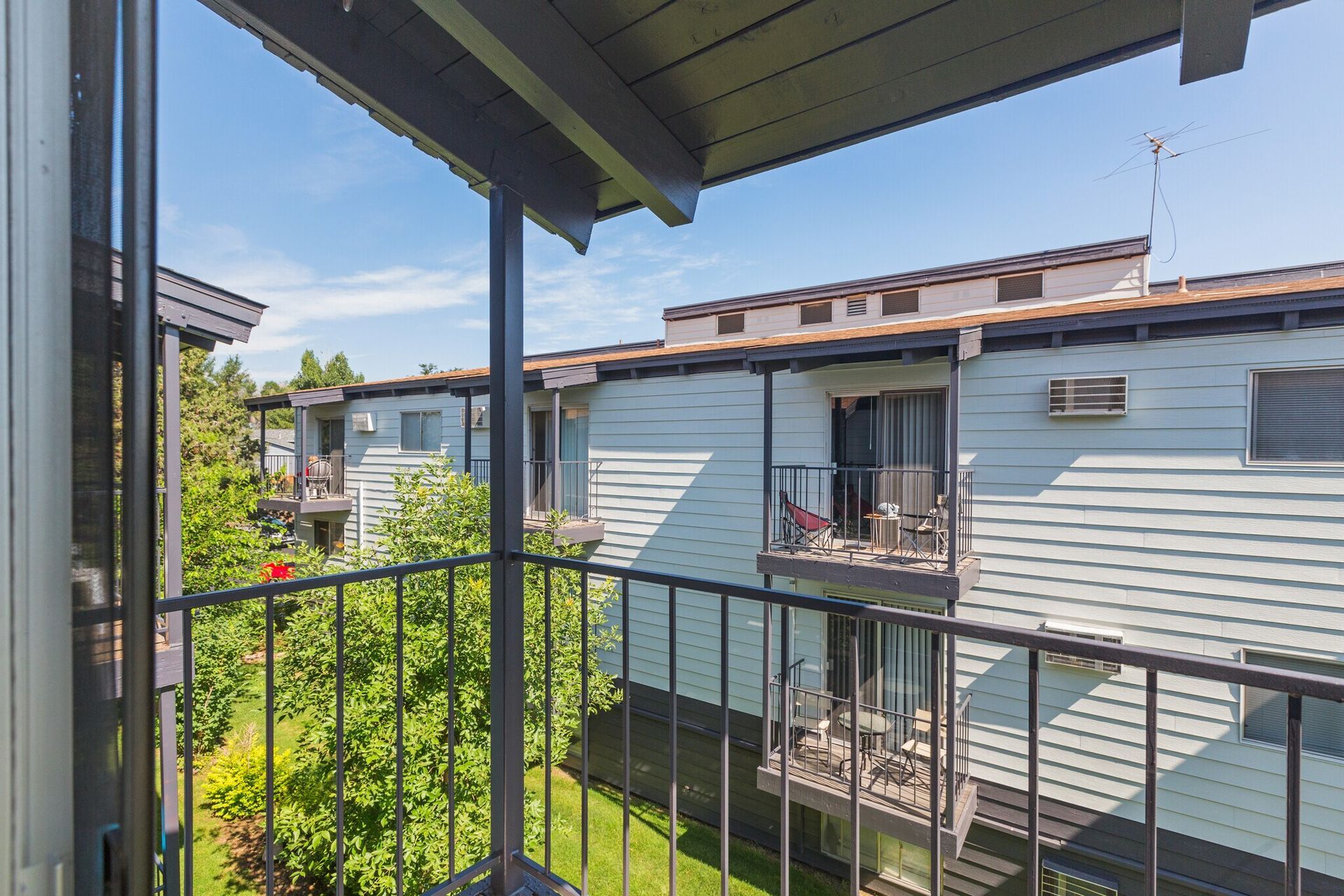 View from an apartment balcony overlooking other apartments with trees and blue sky.