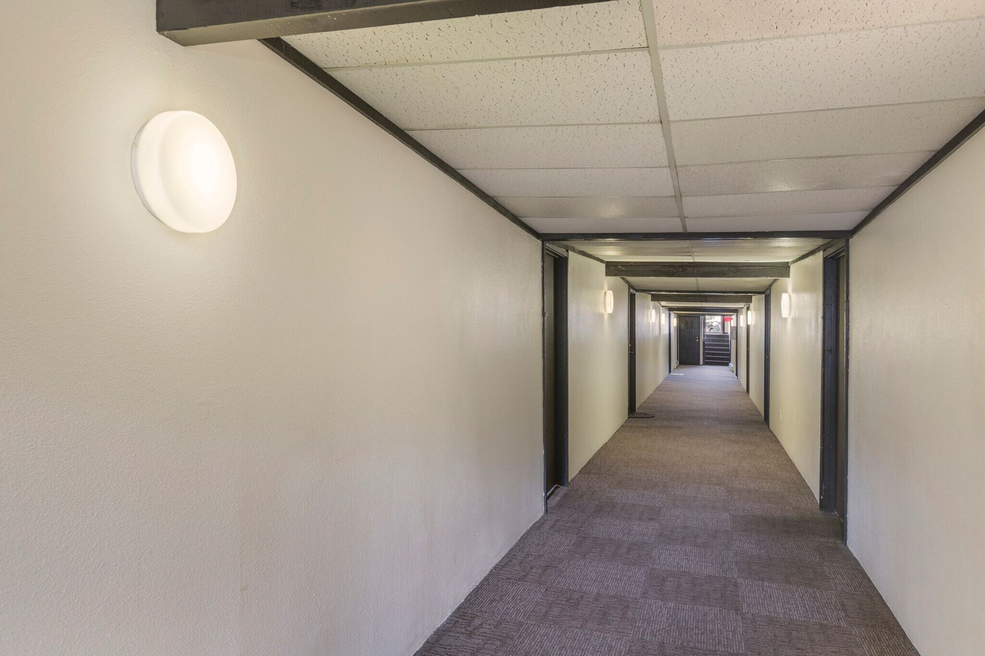 Long, narrow hallway with white walls, recessed ceiling, and dark carpet. Round wall lights illuminate the path.